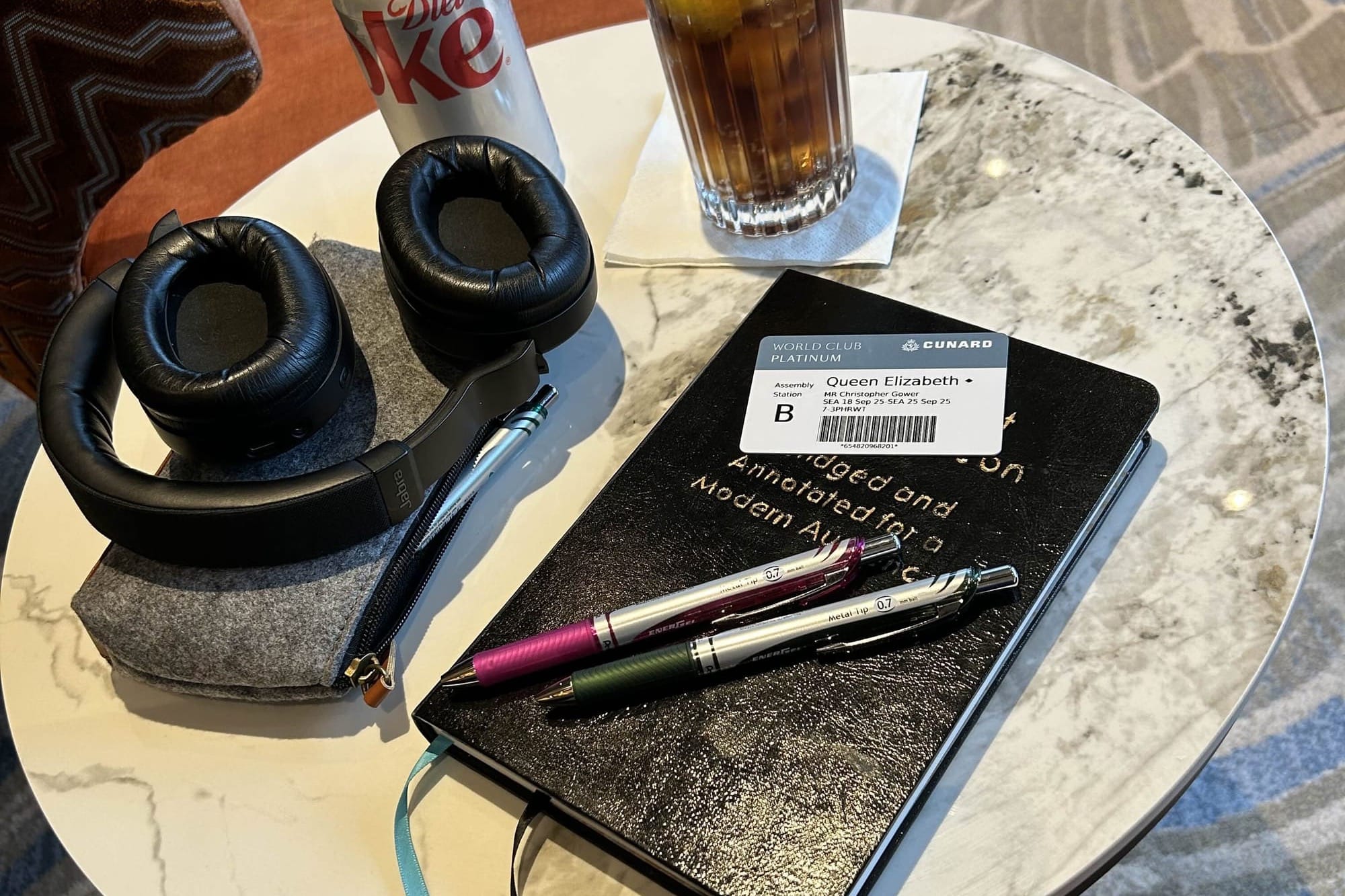 Photo of a round marble cafe tabletop, holding a glass of Diet Coke, a pair of headphones, several pens and a pen holder, a hardcover notebook, and a cruise ship key card.
