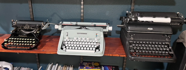 Three typewriters on a shelf. From left to right, a portable 1931 Smith, a green mid-20th century Hermes, and a large 1940’s era Royal.